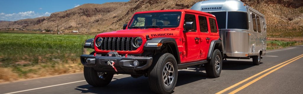 A red Jeep Wrangler Rubicon tows a silver Airstream trailer down a highway. Mountains are in the background.