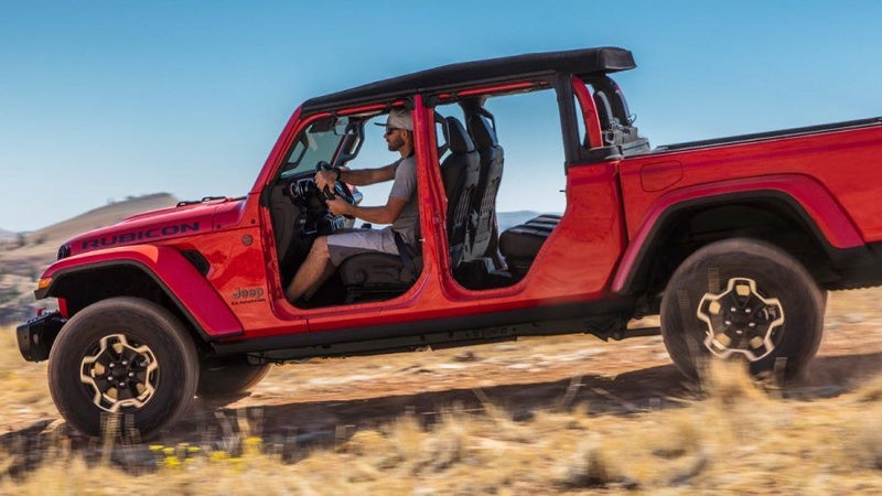 Red Jeep Gladiator Rubicon pickup truck, with doors removed, driving off-road on a sunny day. A man is driving.