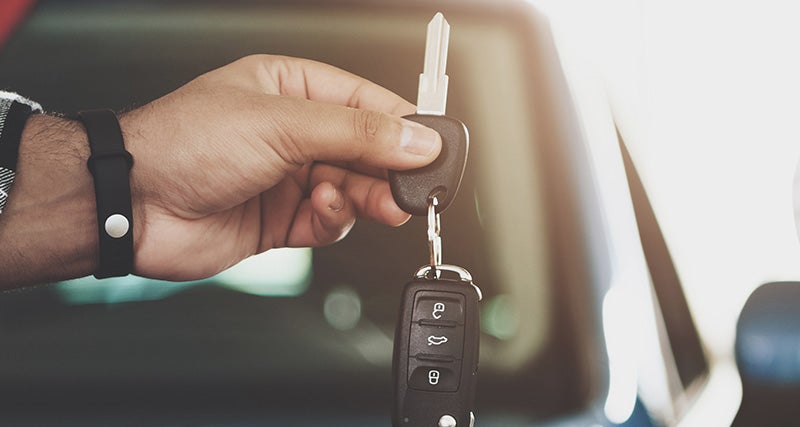 A hand holding a car key and fob, with a blurred car window and bright light in the background.