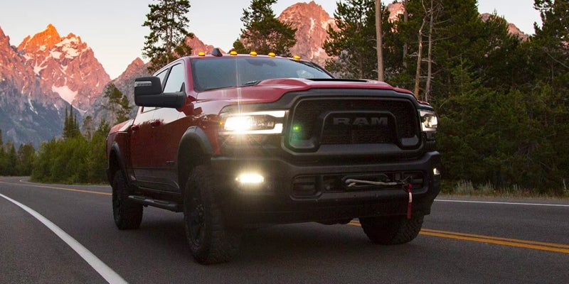 Red Ram truck with bright headlights on a mountain road at dusk. Mountains with an orange glow visible in the background.