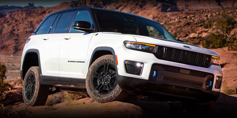 A white Jeep Grand Cherokee, with a black roof, side mirrors, and wheels, on a rocky dirt road. Red rock formations in the background.