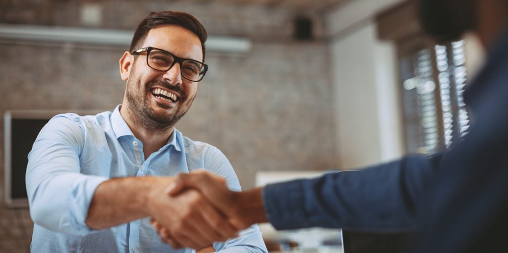 Two men shaking hands in a business setting. The man on the left is smiling and wearing glasses.