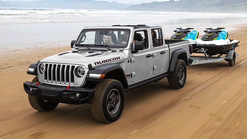 A gray Jeep Gladiator Rubicon pickup drives on a sandy beach, pulling a trailer with two blue and black jet skis.