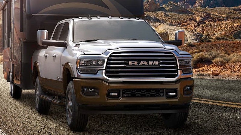 Front view of a white Ram pickup truck with a brown lower body, towing a large camper on a desert road.