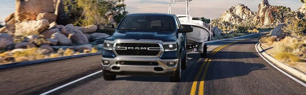 A blue Ram truck tows a boat down a curving highway. Desert rocks and plants line the road.