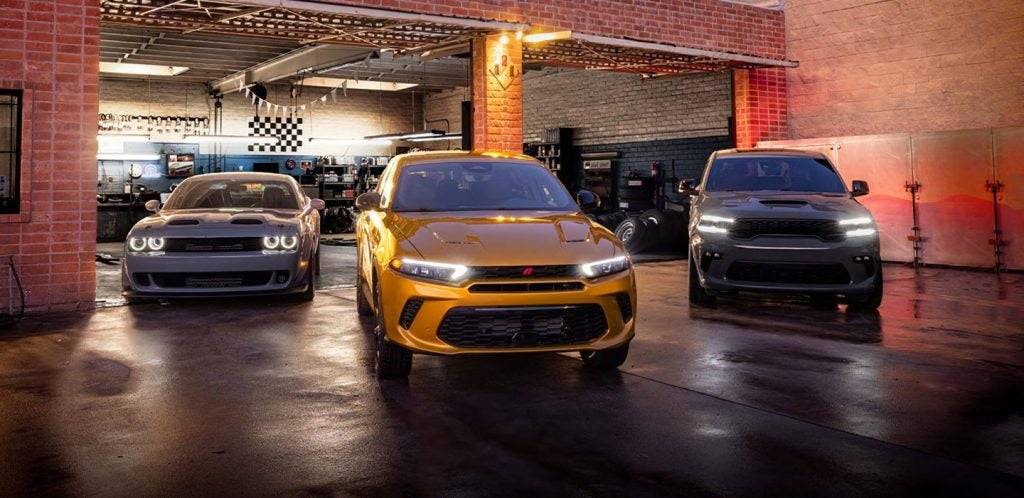 Three Dodge vehicles in a garage: a gray Challenger, golden Hornet, and gray Durango, headlights on.