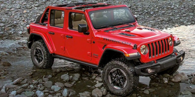 A red Jeep Wrangler Rubicon with its removable top off, parked in a shallow rocky riverbed.