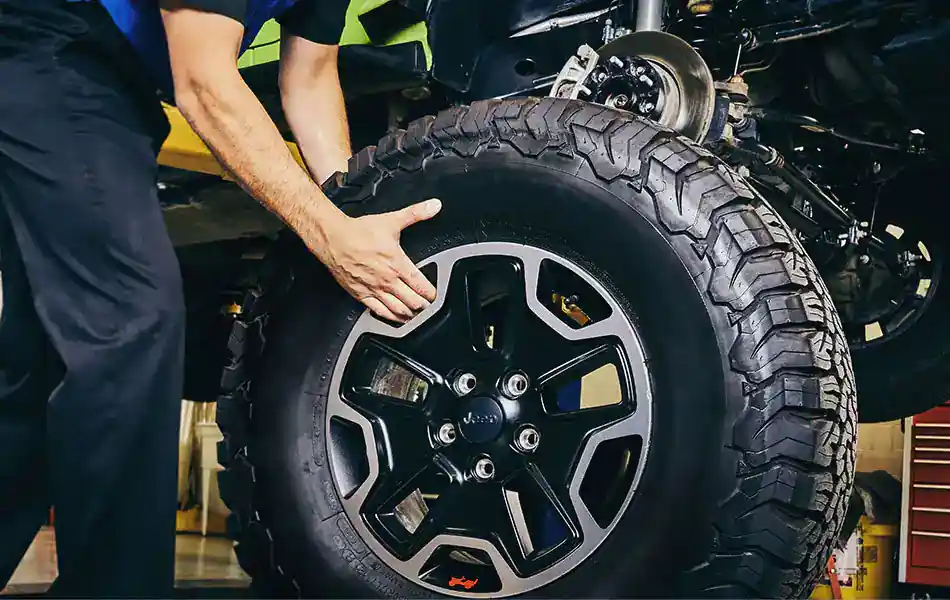 A mechanic installs a large black tire with a black and silver rim onto a vehicle lifted on a hydraulic jack.