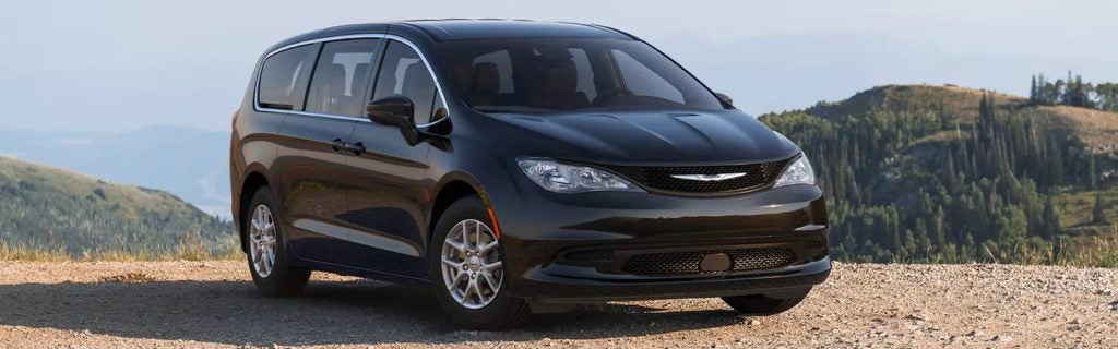 A black minivan is parked on a dirt road, angled slightly towards the right. Mountains and trees are in the background.