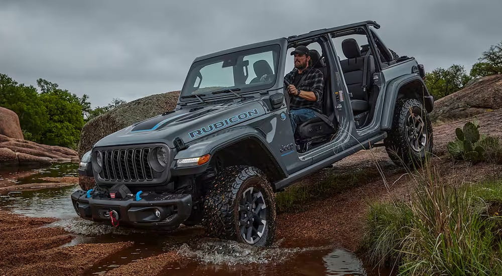 A grey 2024 Jeep Wrangler Rubicon driving off-road across a shallow stream.
