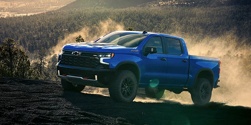 Blue Chevrolet Silverado 1500 driving up slope of mountain framed against forest in distant background.
