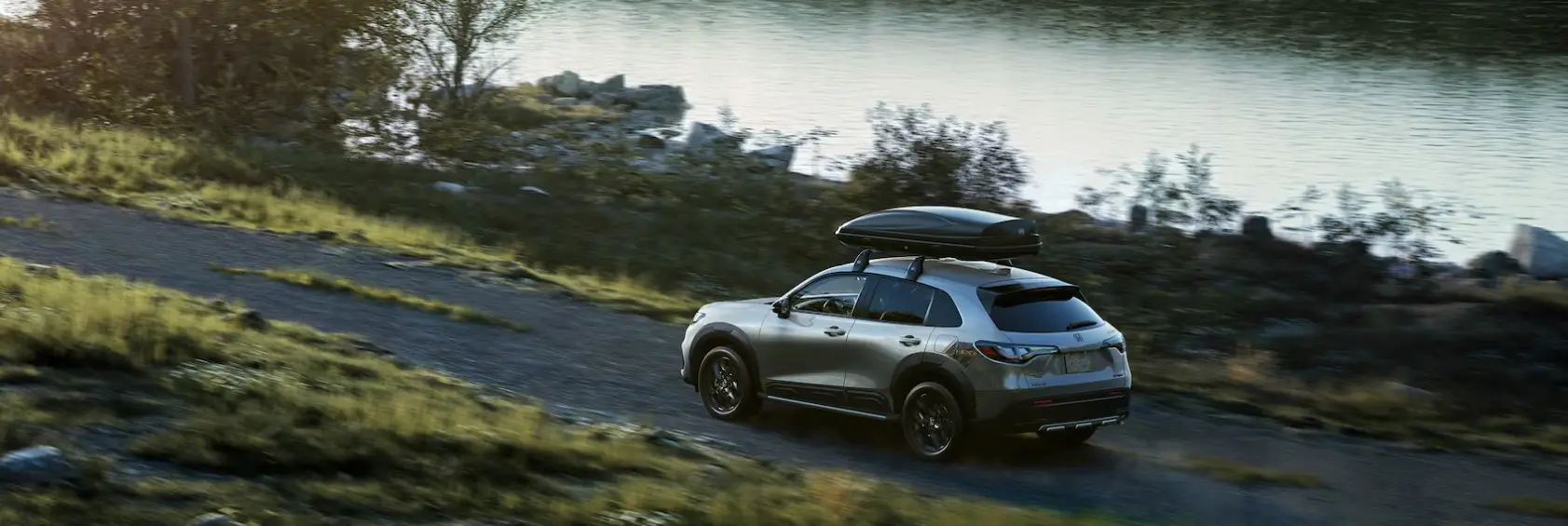 The 2025 Honda HR-V is shown driving on a small road near a lake, making this SUV a great adventurer for you and your family. Shop for your new Honda HR-V at Stonecrest Honda!