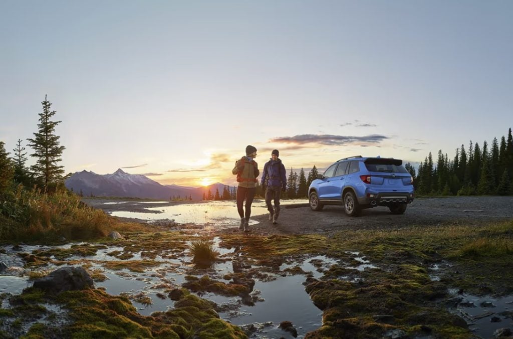 The new 2025 Honda Passport sits parked on off-road terrain with mountains in the background and two people walking away from the car. Shop for your new Honda Passport at Stonecrest Honda.