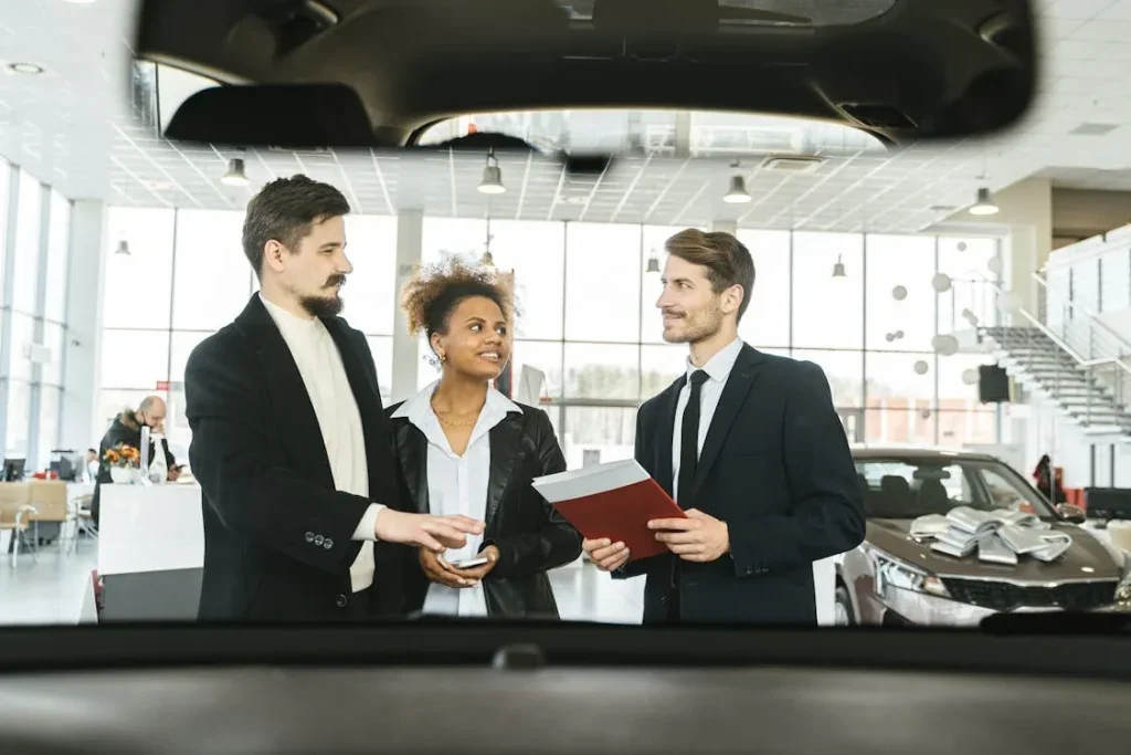 three people standing in a car dealership one man holds a red folder and another man reaches for the folder while having his arm around a shorter woman 