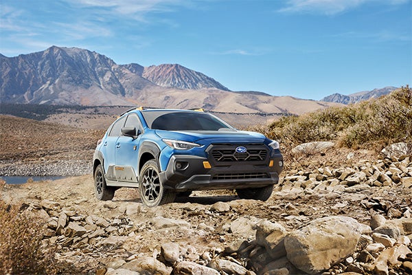 Blue Subaru Crosstrek Wilderness driving along a rocky trail with mountains in the background under a clear sky.