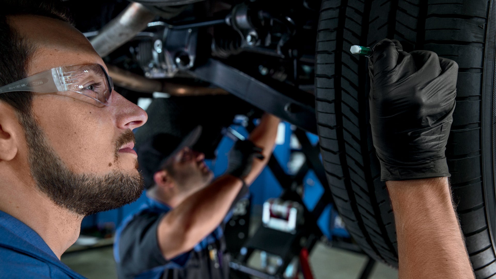Mechanic checks tire tread depth with gauge, wearing safety glasses.