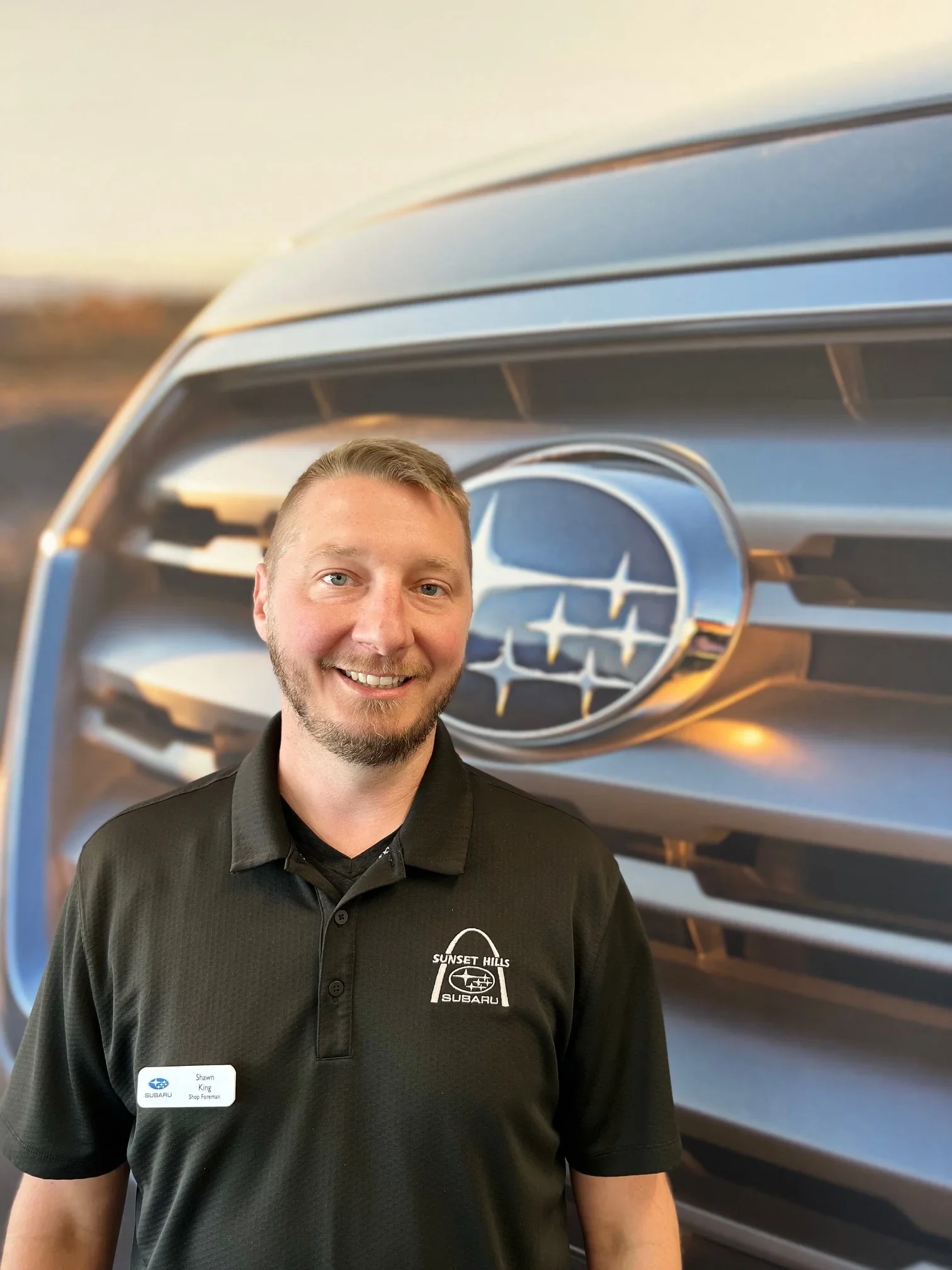 Shawn King, Shop Foreman at Sunset Hills Subaru, smiling in front of a large Subaru logo background.