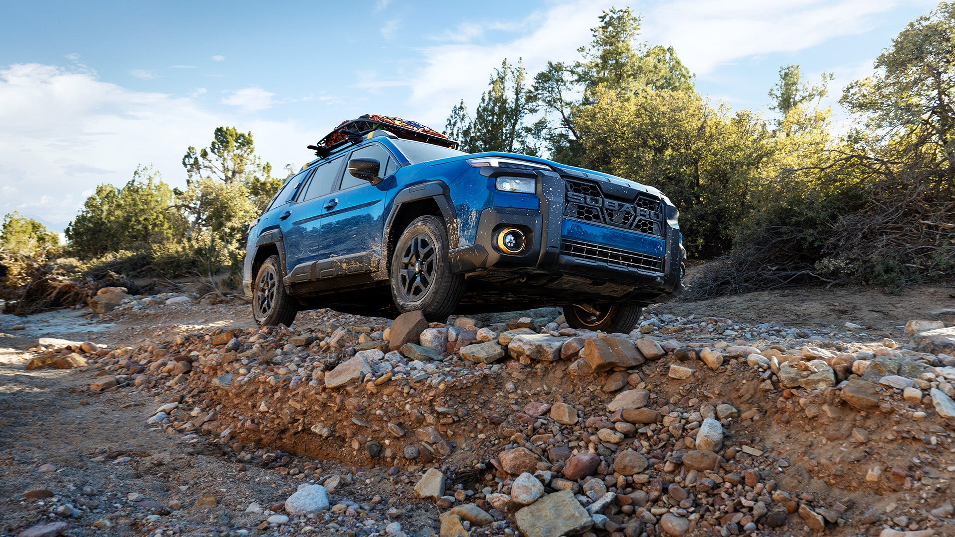 Blue Subaru Forester Wilderness off-roading on a steep, rocky trail under a bright blue sky.