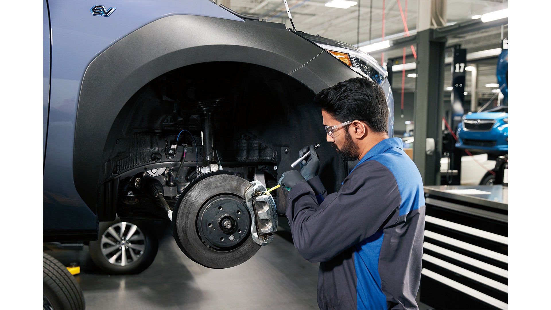 Mechanic in a blue uniform inspecting the brake system of a light blue electric vehicle on a lift.