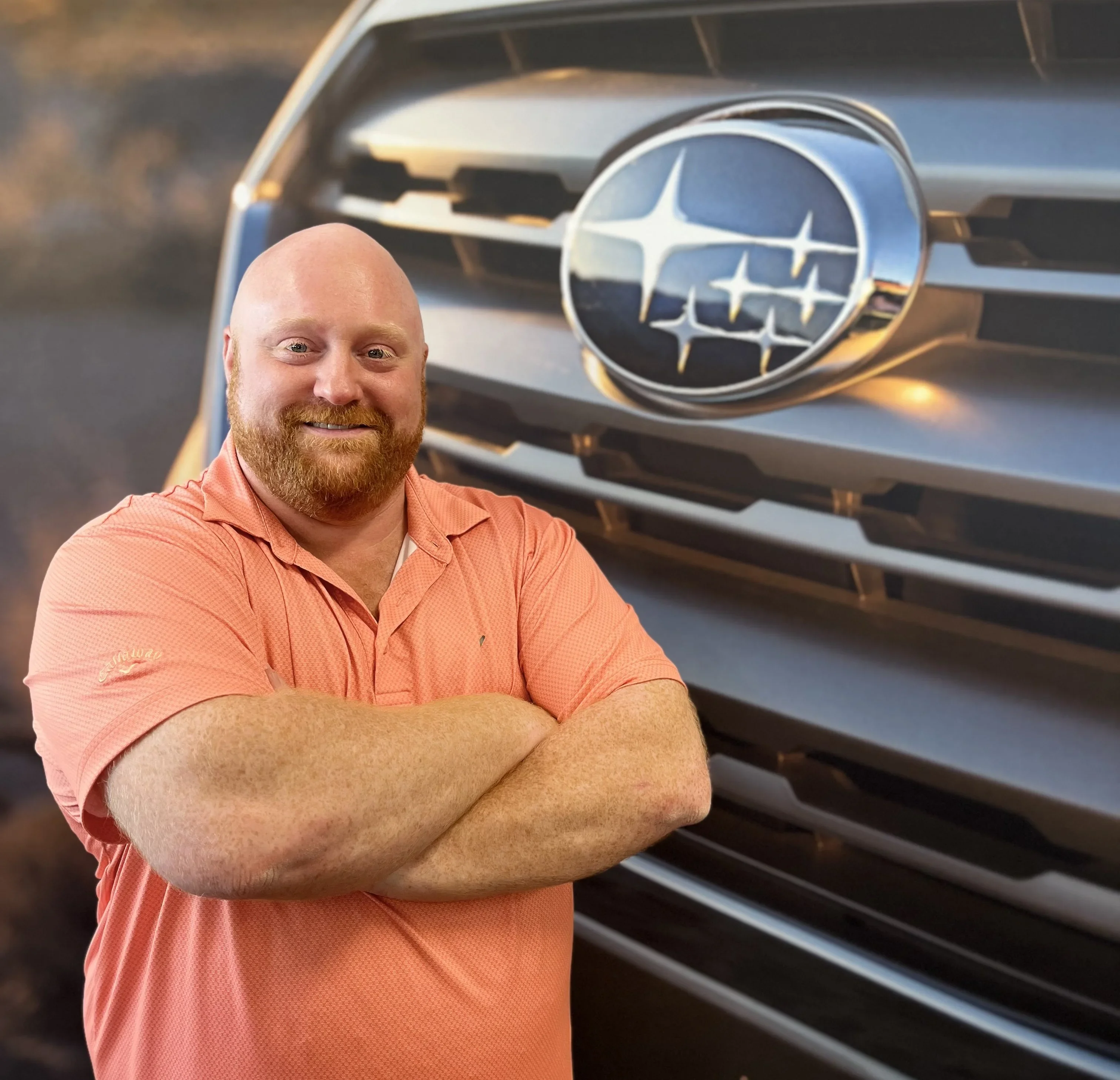 A smiling man with a red beard and orange polo shirt stands with arms crossed in front of a Subaru car grille.