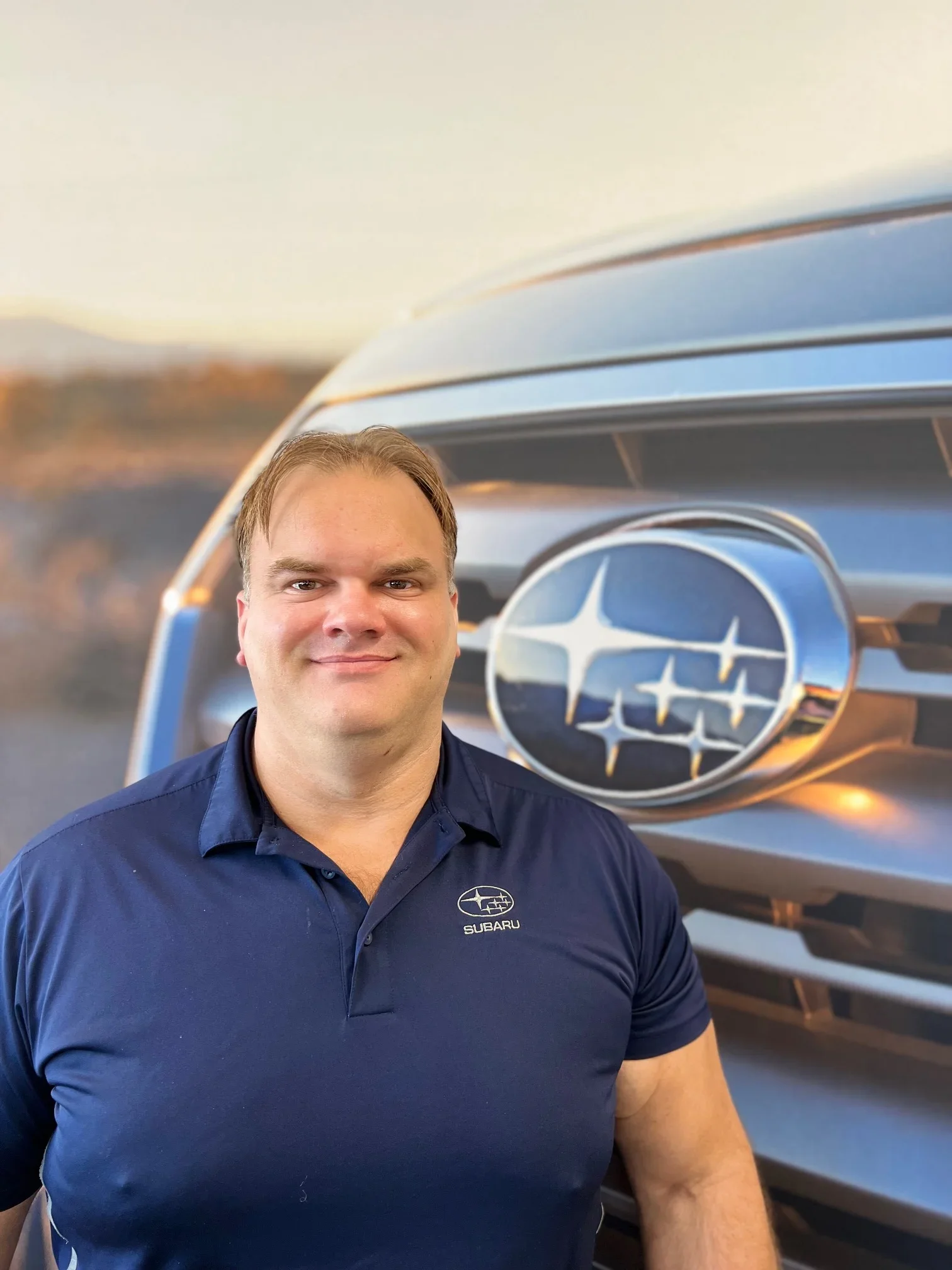 A man in a navy blue Subaru polo shirt smiling in front of a large Subaru logo.
