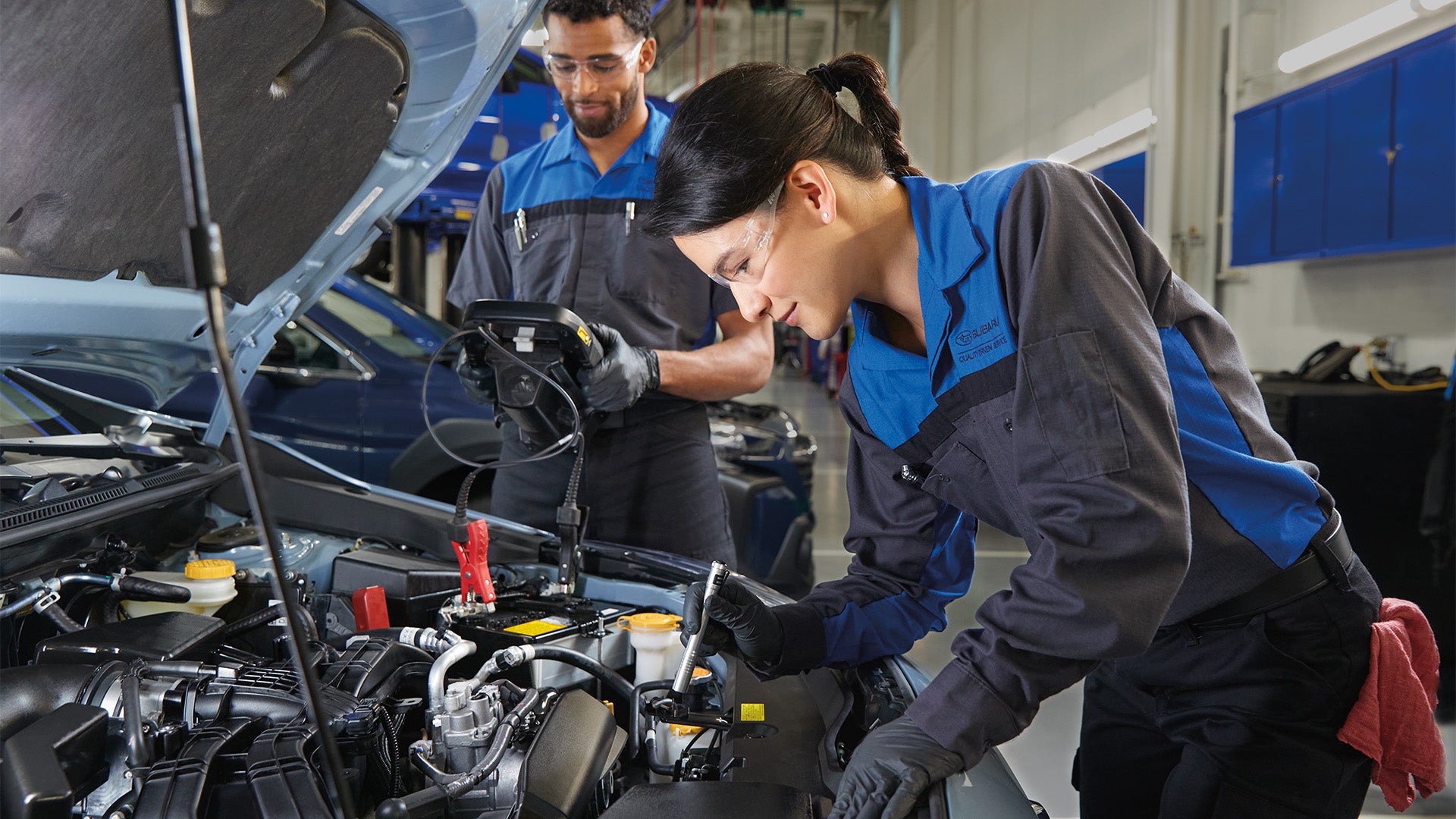 Two Subaru technicians inspecting a car engine with diagnostic tools in a garage.