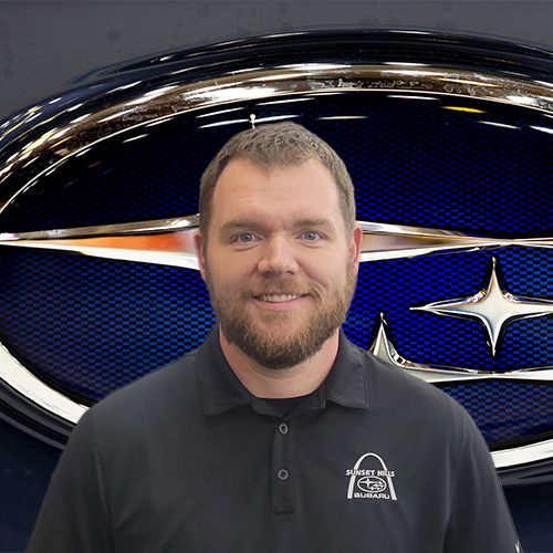 A man with a beard wearing a black Sunset Hills Subaru polo shirt smiling in front of a large Subaru logo.