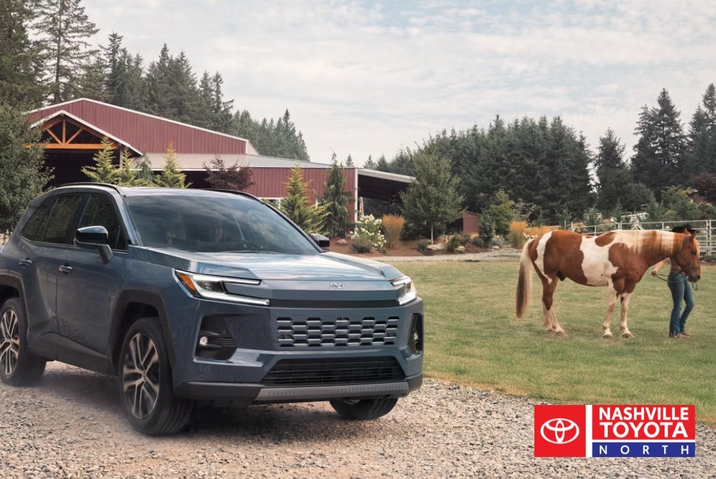 A blue SUV is parked on a gravel road in front of a farm with a red barn, while a person leads a brown and white horse in a grassy field.