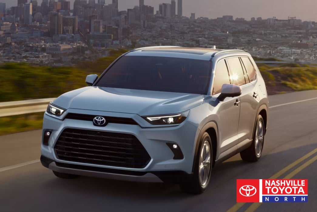 A white Toyota SUV drives on a road with a city skyline in the background during sunset.