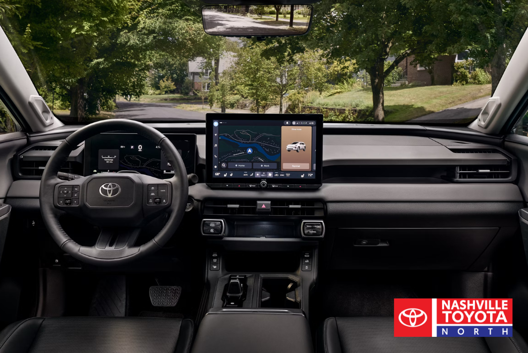 The interior view of a vehicle shows a dashboard with a large touchscreen displaying navigation, a digital instrument cluster, and a steering wheel with controls. Through the windshield, a tree-lined street is visible.