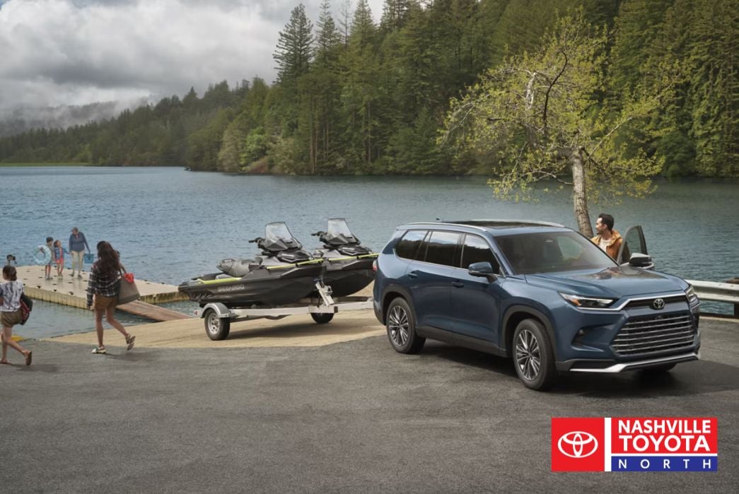 A dark blue SUV is parked next to a trailer carrying two jet skis by a lake with a forested background. People are walking on a dock towards the water.
