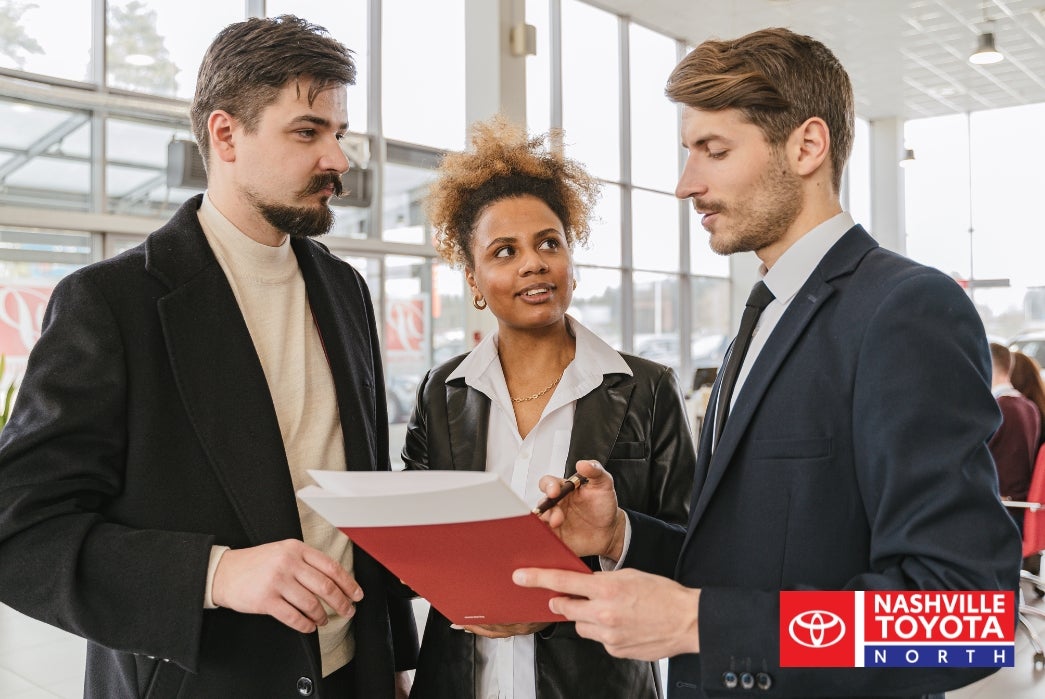 Three people in business attire are looking at a red folder with documents in a dealership.