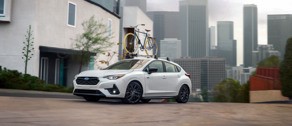 Unlimited Impreza RS with bike rack and bike driving on wet road, cityscape in background.
