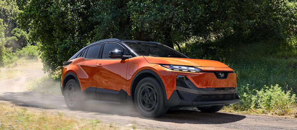 Orange Subaru SUV driving on a dirt road surrounded by greenery and trees.
