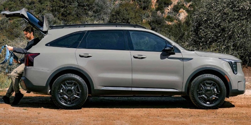 A man resting against the open trunk of a grey 2025 Kia Sorento parked on rugged terrain on a bright, sunny day