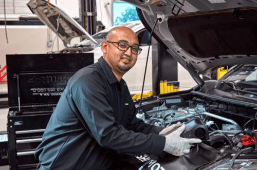 A mechanic wearing glasses and gloves smiles while working on a car engine in a garage. The open hood and toolbox indicate a repair in progress.