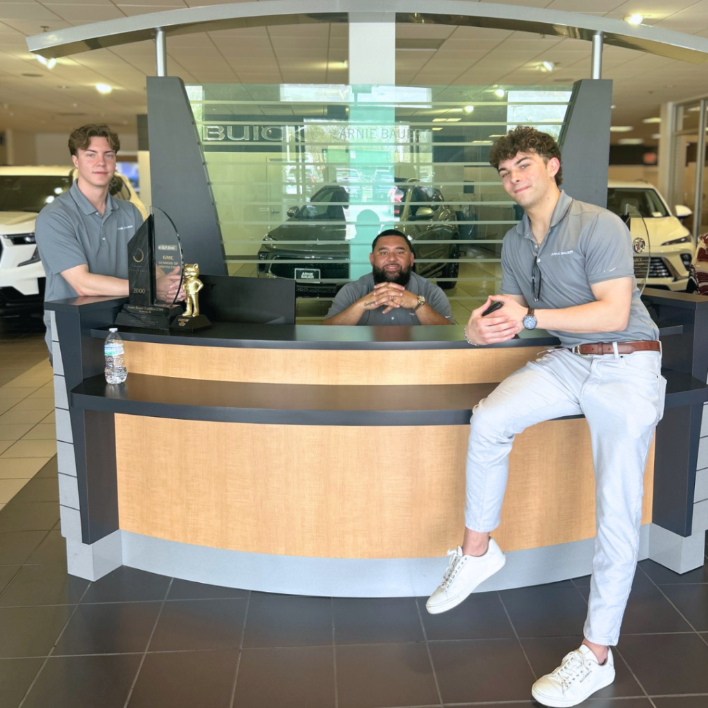 Three professional sales representatives smiling at the Arnie Bauer Buick GMC welcome desk in Matteson, IL, showcasing the human customer service and personal accountability behind our remote online car buying experience.