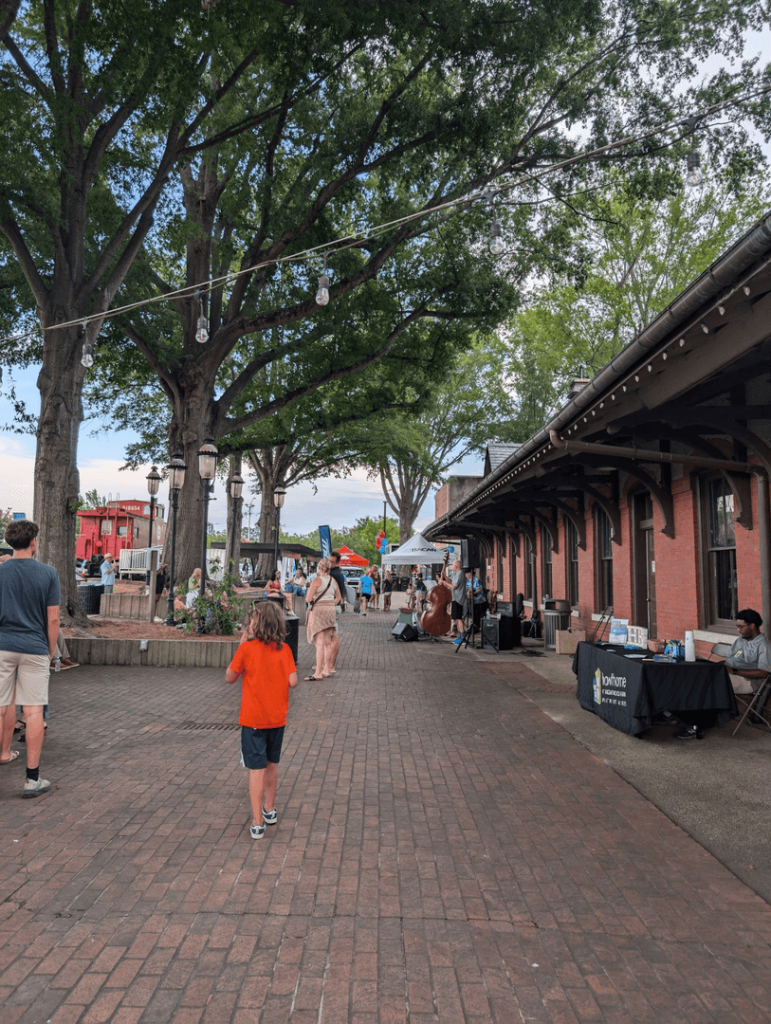 People walking along a tree-lined brick walkway with string lights and market stalls beside a red brick building.