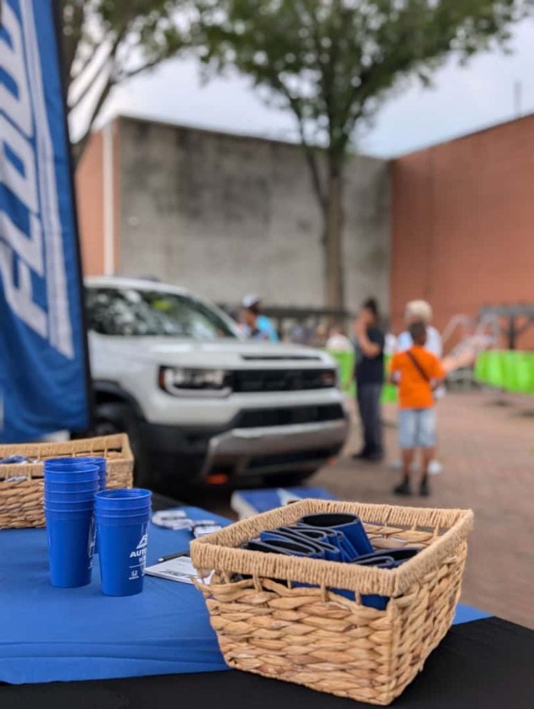 Promotional event table with blue cups and branded items in a basket, set up in front of a white SUV, with a few people talking in the background outdoors.