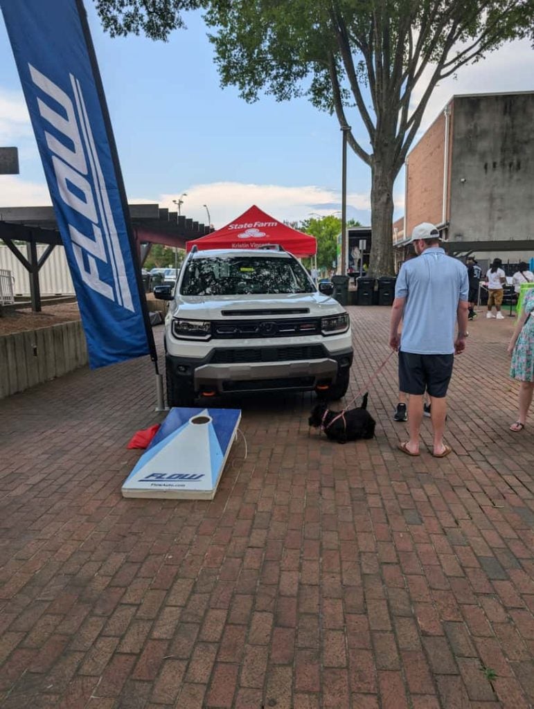 White pickup truck at an outdoor event with a cornhole game, branded flags, and people walking a small dog on a brick plaza.