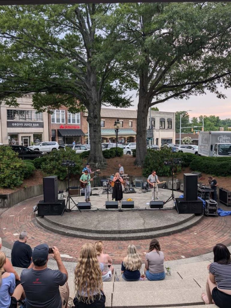 Live band performing on an outdoor stage in a small park, with seated audience and storefronts in the background.