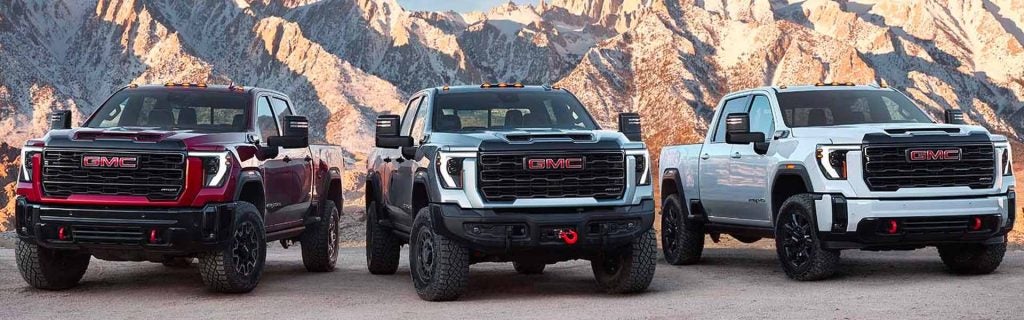 Lineup of 2025 GMC Sierra HD trucks parked outdoors with mountain backdrop showcasing rugged design and off-road capability.