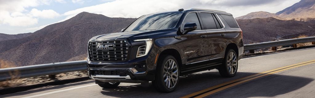 Black 2026 GMC Yukon Denali SUV driving on a mountain road with desert hills in the background.