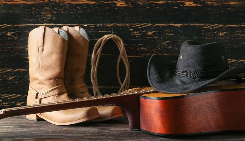 beige cowboy boots sitting next to a guitar that a cowboy hat has been perched on top of 