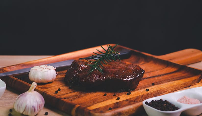 a steak lays on a cutting board and a sprig of rosemary is placed on top of it with heads of garlic surrounding it 