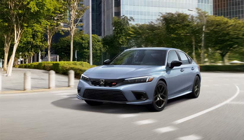 a silver honda drives down a road that has trees and bushes planted along the sidewalk with a business building in the background 