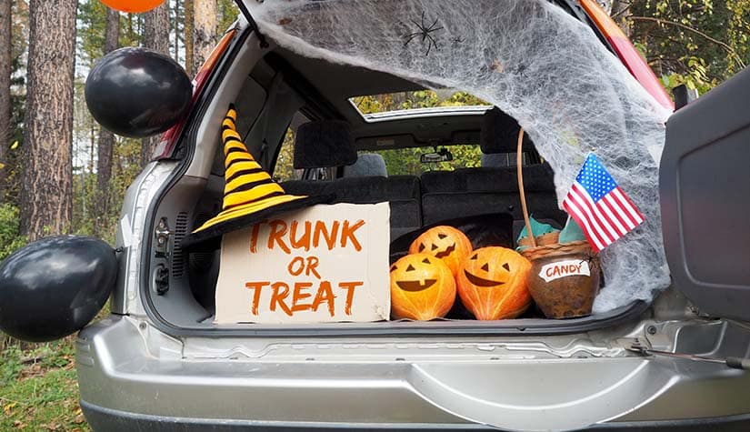 a grey minivan has the back trunk door open. its decorated with black balloons, pumpkins, and a witch hat on top of a sign that says trick or treat 