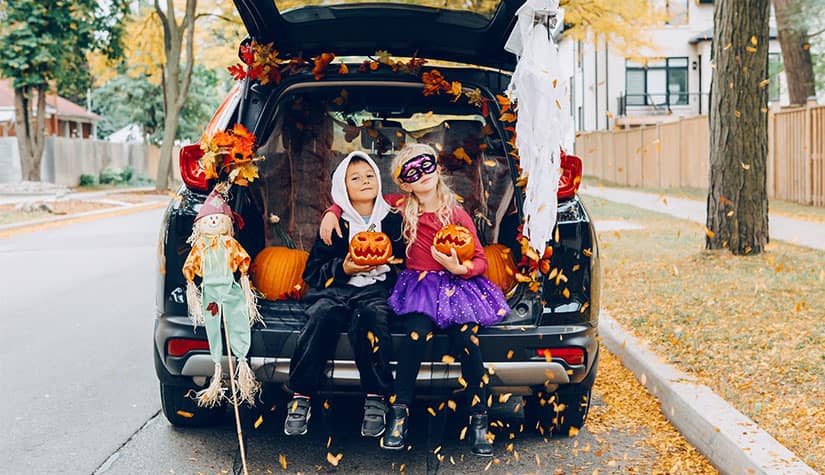 two small children in halloween costumes sit next to each other in the open trunk of an SUV