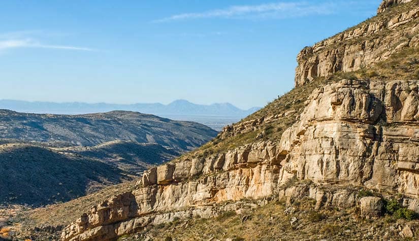 a craggy mountainside sits in the foreground with hills and a blue sky in the background 