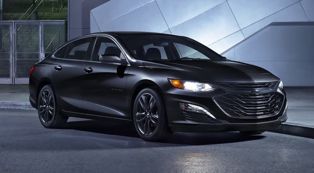 A black 2024 Chevy Malibu Midnight parked outside a dealer with used cars for sale.